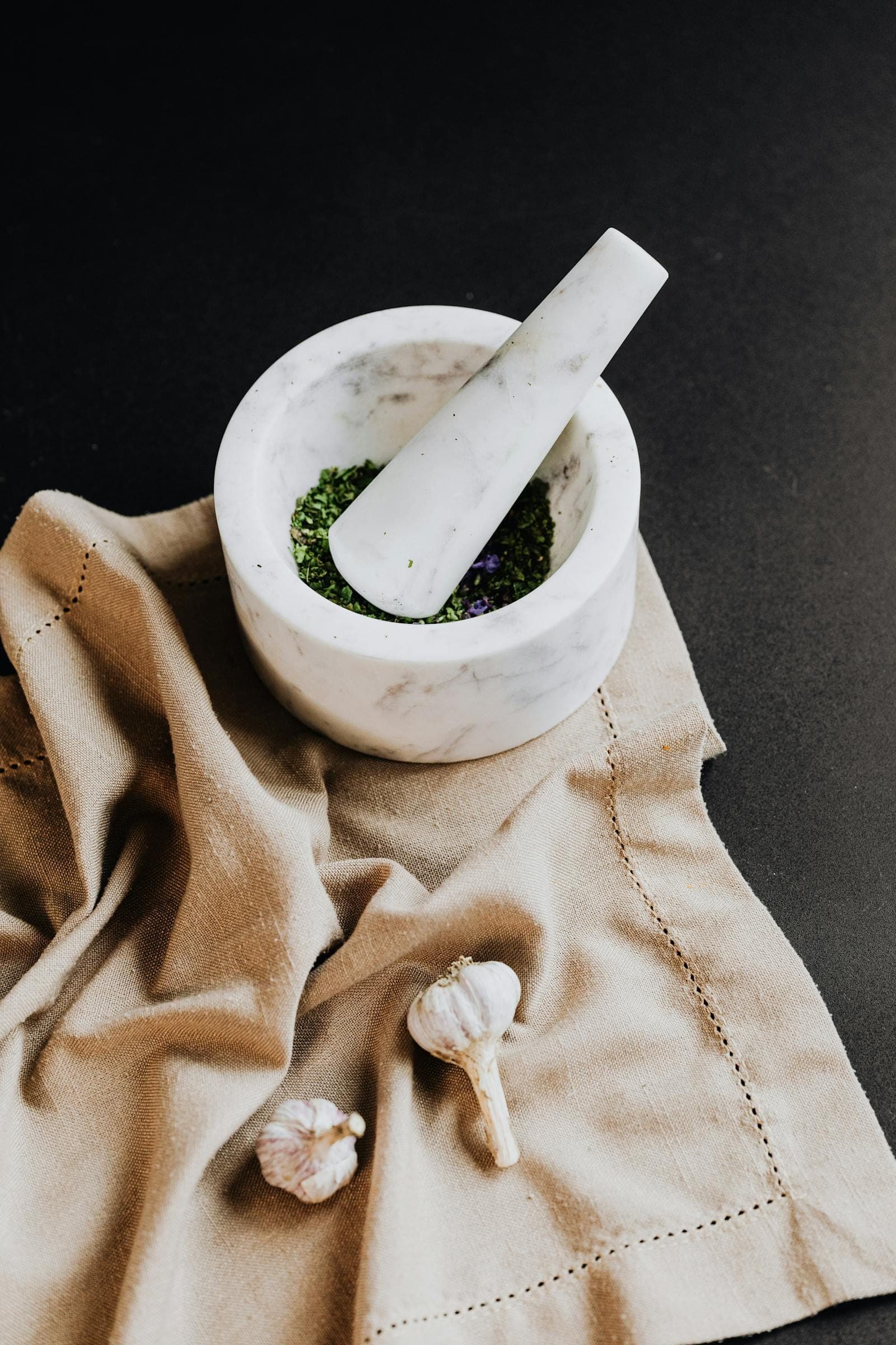 White marble mortar with fresh herbs being ground, on warm linen, with garlic bulbs